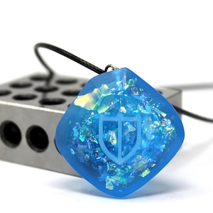 Puede incluir: Un colgante cuadrado azul con purpurina iridiscente incrustada en la resina. El colgante tiene un dise&ntilde;o de escudo grabado en la superficie y est&aacute; unido a un cord&oacute;n negro.