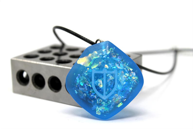 Puede incluir: Un colgante cuadrado azul con purpurina iridiscente incrustada en la resina. El colgante tiene un dise&ntilde;o de escudo grabado en la superficie y est&aacute; unido a un cord&oacute;n negro.