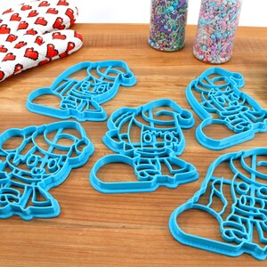 May include: Five blue plastic cookie cutters shaped like elves wearing Santa hats. The cutters are on a wooden surface.