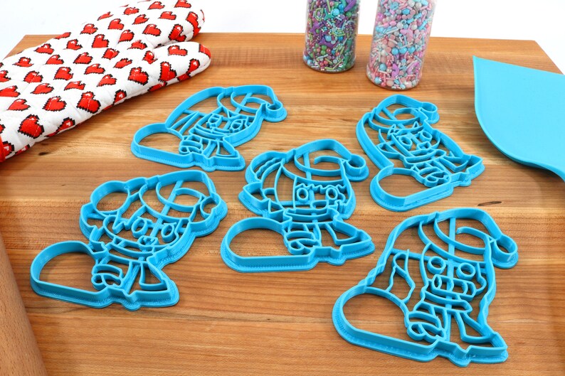 May include: Five blue plastic cookie cutters shaped like elves wearing Santa hats. The cutters are on a wooden surface.