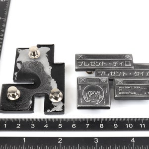 Op de afbeelding: Twee zilveren metalen pinnen met een zwart-wit ontwerp. De pinnen zijn gevormd als computervensters met tekst die "プレゼント・ディロ" en "プレゼント・タイムロ" luidt. Het onderste venster heeft een cartoon gezicht met de tekst "YOU DONT SEEM, TO UNDERSTAND... A SHAME".
