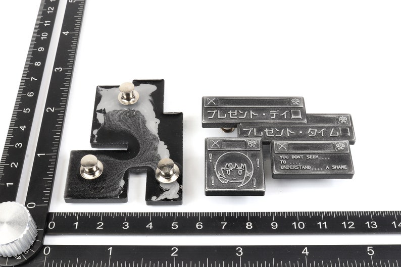 Op de afbeelding: Twee zilveren metalen pinnen met een zwart-wit ontwerp. De pinnen zijn gevormd als computervensters met tekst die "プレゼント・ディロ" en "プレゼント・タイムロ" luidt. Het onderste venster heeft een cartoon gezicht met de tekst "YOU DONT SEEM, TO UNDERSTAND... A SHAME".