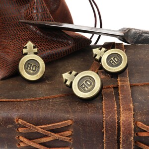 May include: Three gold-colored metal tokens with a symbol and the letters "AD" on a brown leather journal with a braided leather strap.