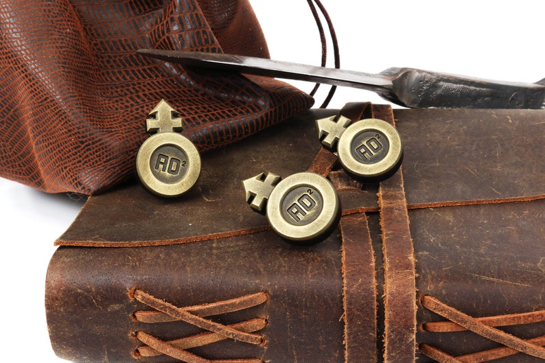 May include: Three gold-colored metal tokens with a symbol and the letters "AD" on a brown leather journal with a braided leather strap.