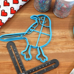 Puede incluir: Un cortador de galletas de pl&aacute;stico azul con forma de arrendajo azul. El cortador est&aacute; sobre una superficie de madera con una regla y una esp&aacute;tula.
