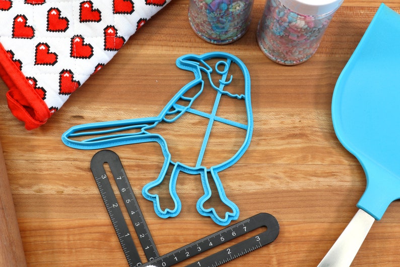 Puede incluir: Un cortador de galletas de pl&aacute;stico azul con forma de arrendajo azul. El cortador est&aacute; sobre una superficie de madera con una regla y una esp&aacute;tula.