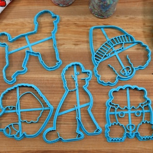 May include: Five blue plastic cookie cutters shaped like poodles. The cutters are on a wooden surface.