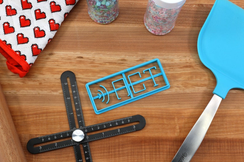 May include: A blue plastic cookie cutter shaped like the word "ACT" on a wooden surface with a black ruler, a blue spatula, and two jars of sprinkles.