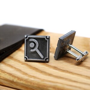 May include: A pair of silver-toned cufflinks with a square design and a magnifying glass symbol. The cufflinks are displayed on a wooden surface next to a brown leather case with a metal button.