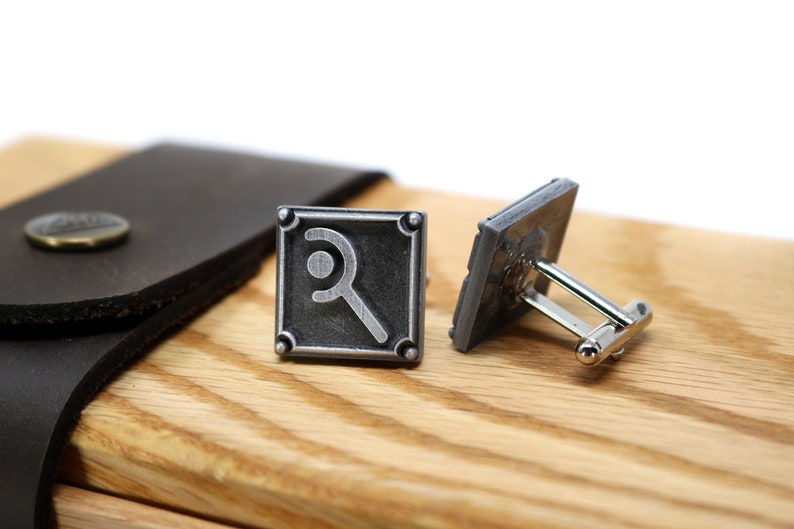 May include: A pair of silver-toned cufflinks with a square design and a magnifying glass symbol. The cufflinks are displayed on a wooden surface next to a brown leather case with a metal button.