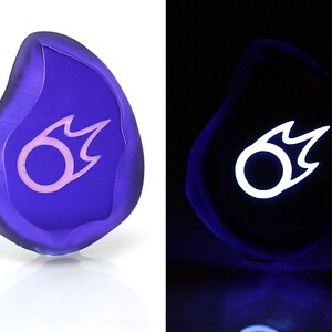 May include: A purple glass stone with a white and pink flame symbol. The stone glows white under a blacklight.