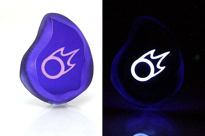 May include: A purple glass stone with a white and pink flame symbol. The stone glows white under a blacklight.