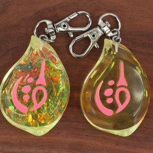 May include: Two teardrop-shaped keychains with a pink symbol inside. One keychain is clear yellow and the other is iridescent.