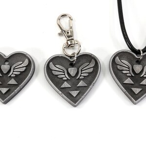 Puede incluir: Tres colgantes de coraz&oacute;n plateados con un coraz&oacute;n alado y un dise&ntilde;o triangular. Un colgante est&aacute; unido a un llavero plateado, otro est&aacute; unido a un cord&oacute;n negro y el tercero es un colgante independiente.