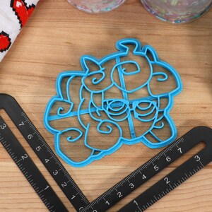 May include: A blue plastic cookie cutter shaped like a cartoon unicorn with a horn, mane, and tail. The cutter is on a wooden surface with a ruler.