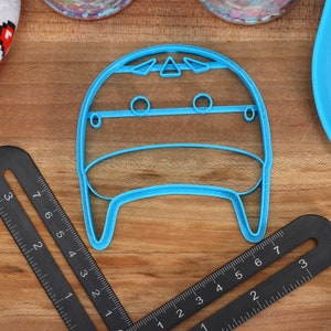 May include: A light blue plastic cookie cutter shaped like a cartoon character with a round head, two eyes, and a mouth. The cookie cutter is on a wooden surface.