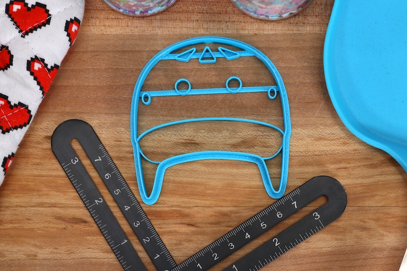 May include: A light blue plastic cookie cutter shaped like a cartoon character with a round head, two eyes, and a mouth. The cookie cutter is on a wooden surface.