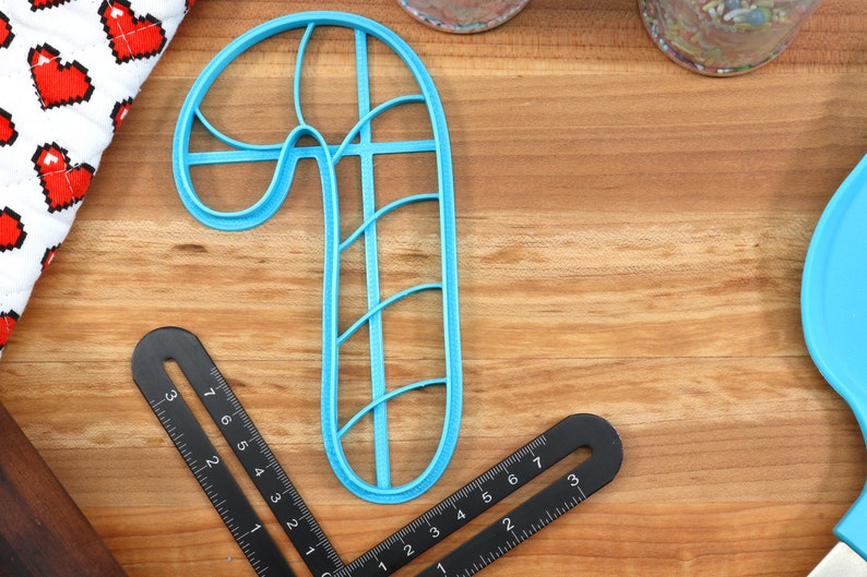 Op de afbeelding: Een blauwe plastic koekjesvormer in de vorm van een snoepstok op een houten oppervlak. De koekjesvormer heeft een gedetailleerd ontwerp met lijnen en bochten.