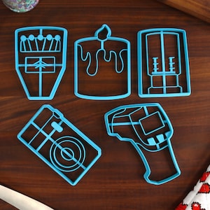 May include: A collection of five blue plastic cookie cutters in various shapes, including a candle, a matchbox, and a thermometer. The cutters are arranged on a dark wooden surface, with sprinkles and a spatula visible in the background.
