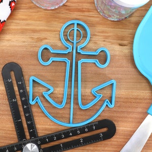 May include: A light blue anchor-shaped cookie cutter sits on a wooden surface, next to a black angle ruler and a blue spatula. A white cloth with red pixelated hearts is in the background. The ruler has measurements in inches.