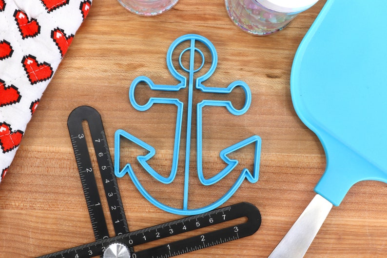 May include: A light blue anchor-shaped cookie cutter sits on a wooden surface, next to a black angle ruler and a blue spatula. A white cloth with red pixelated hearts is in the background. The ruler has measurements in inches.