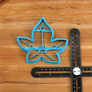 May include: A blue plastic leaf-shaped cookie cutter with a detailed design. The cutter is on a wooden surface next to a black ruler.