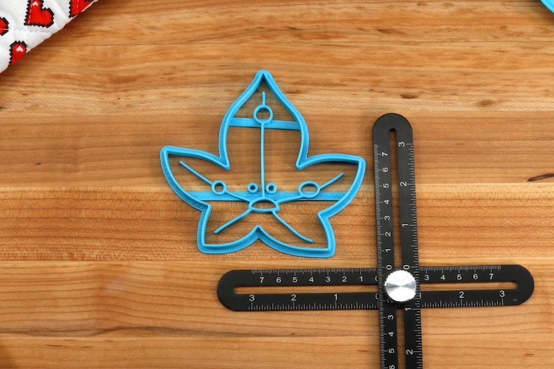 May include: A blue plastic leaf-shaped cookie cutter with a detailed design. The cutter is on a wooden surface next to a black ruler.