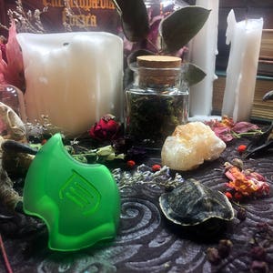 May include: A green glass pendant with a stylized design, surrounded by dried flowers, a small jar of herbs, a white candle, and a white crystal on a dark brown surface.
