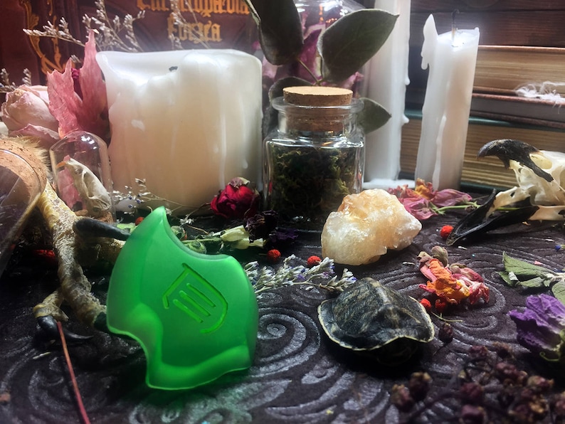 May include: A green glass pendant with a stylized design, surrounded by dried flowers, a small jar of herbs, a white candle, and a white crystal on a dark brown surface.