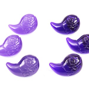 K&ouml;nnte beinhalten: Sechs violette, geisterf&ouml;rmige Perlen mit einem erhabenen Design. Die Perlen sind aus Glas oder Kunststoff und haben eine gl&auml;nzende Oberfl&auml;che.