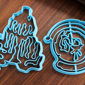 Puede incluir: Dos cortadores de galletas de pl&aacute;stico azul, uno con forma de mano y el otro con forma de c&iacute;rculo con un dise&ntilde;o en su interior.