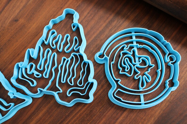 Puede incluir: Dos cortadores de galletas de pl&aacute;stico azul, uno con forma de mano y el otro con forma de c&iacute;rculo con un dise&ntilde;o en su interior.