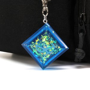 May include: A blue diamond-shaped keychain with iridescent glitter and a silver chain. The keychain has a white design inside.
