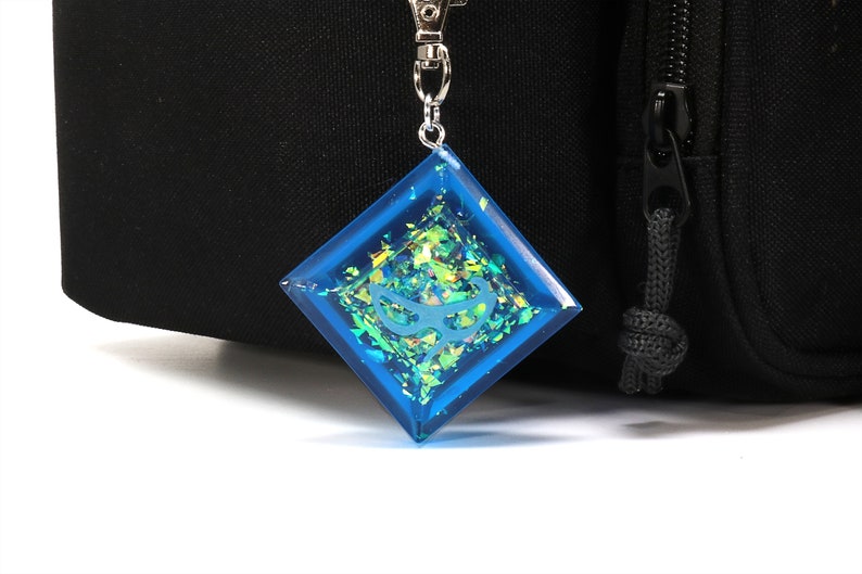 May include: A blue diamond-shaped keychain with iridescent glitter and a silver chain. The keychain has a white design inside.