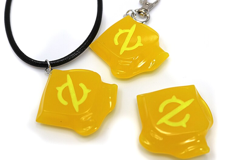 May include: Three yellow plastic charms with a stylized letter Z design. One charm is attached to a black cord necklace, and another charm is attached to a silver key chain.