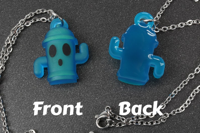 May include: A blue translucent necklace with a cactus-shaped pendant. The front of the pendant has a cartoon face with black eyes and a surprised expression. The back of the pendant is plain. The necklace is made of a silver chain.