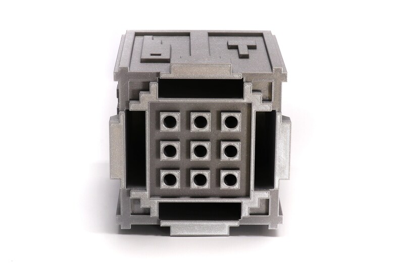 May include: A silver metal box with a grid of nine round holes on the front. The box has a textured surface and appears to be made of a 3D printed material.