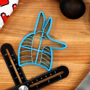 May include: A blue plastic cookie cutter shaped like an Anubis head, a jackal-headed god in ancient Egyptian mythology. The cutter is on a wooden surface with a ruler and other kitchen tools.