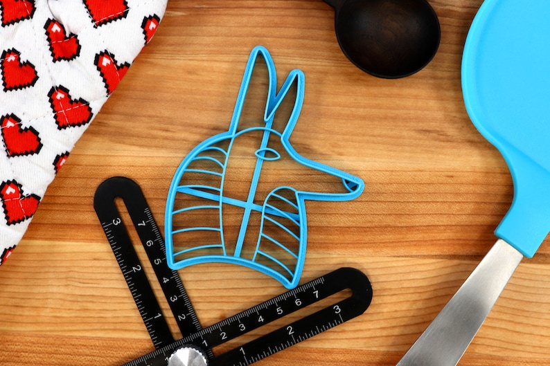 May include: A blue plastic cookie cutter shaped like an Anubis head, a jackal-headed god in ancient Egyptian mythology. The cutter is on a wooden surface with a ruler and other kitchen tools.