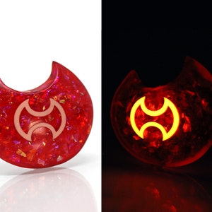 May include: A red crescent moon-shaped resin pendant with a white symbol inside. The pendant glows in the dark, revealing a yellow symbol.
