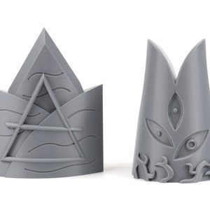 May include: Two gray 3D printed crown-shaped objects. The first crown has a triangular design with wavy lines and a horizontal line across the center. The second crown has a pointed top with a stylized eye design.