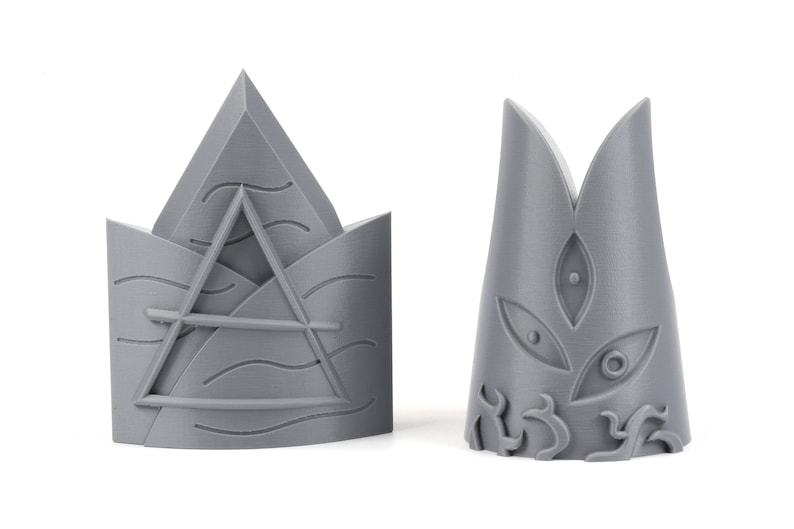 May include: Two gray 3D printed crown-shaped objects. The first crown has a triangular design with wavy lines and a horizontal line across the center. The second crown has a pointed top with a stylized eye design.