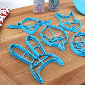 May include: A set of six blue plastic cookie cutters shaped like a bunny, an alien, a bird, a fox, a monster, and an egg. The cutters are on a wooden surface.