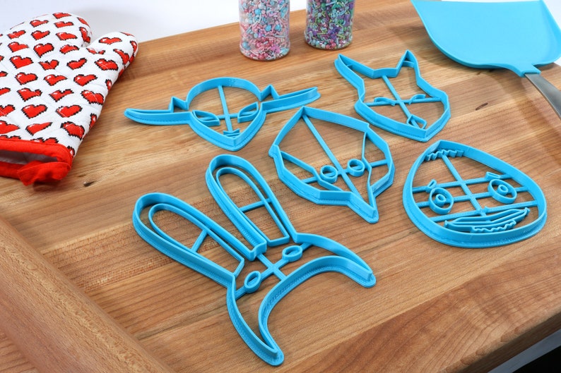 May include: A set of six blue plastic cookie cutters shaped like a bunny, an alien, a bird, a fox, a monster, and an egg. The cutters are on a wooden surface.