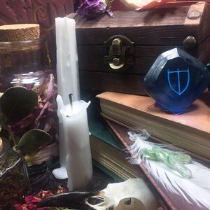 May include: A blue glass gem with a silver shield design sits on a stack of books. Two white candles, a glass jar filled with dried flowers and leaves, and a wooden box with a metal latch are also in the image.