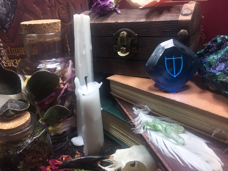 May include: A blue glass gem with a silver shield design sits on a stack of books. Two white candles, a glass jar filled with dried flowers and leaves, and a wooden box with a metal latch are also in the image.