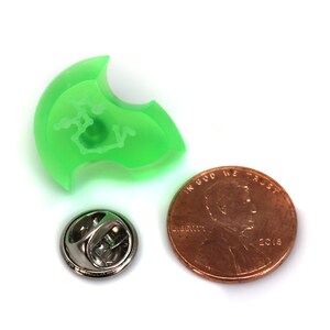 May include: A green plastic pin with a white design. The pin is next to a US penny for size comparison.