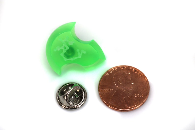 May include: A green plastic pin with a white design. The pin is next to a US penny for size comparison.