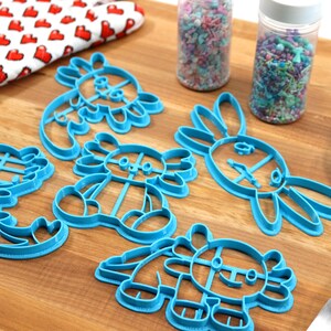 May include: A set of six blue plastic cookie cutters shaped like different animals, including a dragon, a bunny, and a dinosaur. The cutters are on a wooden surface with sprinkles in jars and a blue spatula.