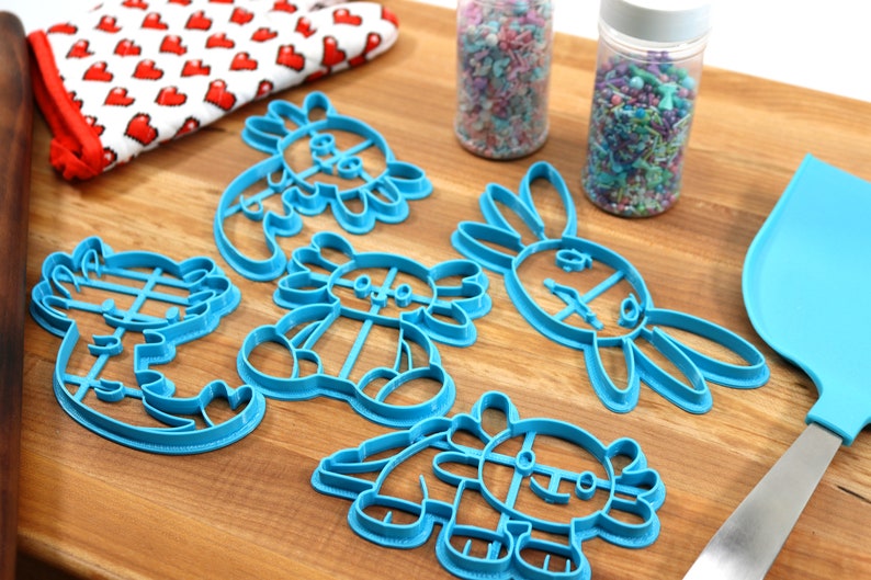 May include: A set of six blue plastic cookie cutters shaped like different animals, including a dragon, a bunny, and a dinosaur. The cutters are on a wooden surface with sprinkles in jars and a blue spatula.